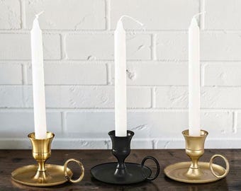 May include: Three metal candle holders with circular bases and side ring handles. Two holders have a brushed gold finish, and one central holder has a matte black finish. Each holder contains a tall, slender, white taper candle standing upright.