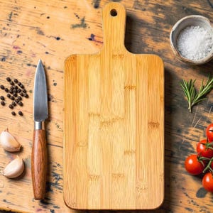 Puede incluir: Una tabla de cortar rectangular de bambú con un mango redondeado y un orificio para colgar. Junto a ella hay un cuchillo de cocina con mango de madera, granos de pimienta negra, dos dientes de ajo, un cuenco con sal y romero fresco.