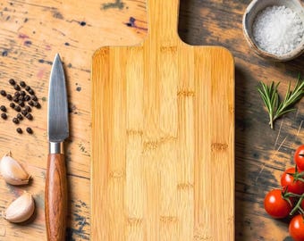 May include: A rectangular bamboo chopping board with a rounded handle and a hole for hanging. Beside it lies a paring knife with a wooden handle, scattered black peppercorns, two garlic cloves, a small bowl of sea salt, and fresh rosemary sprigs.