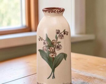 May include: A cylindrical ceramic vase with a cream-coloured base and a narrow neck. The vase features a botanical illustration of lily of the valley flowers with green leaves and white bell-shaped blossoms. The rim and base have a brown speckled trim.