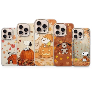 May include: Five clear smartphone cases featuring Snoopy and Woodstock in autumn-themed designs. The cases display illustrations of Snoopy on a pumpkin, inside a jack-o'-lantern, playing in fallen leaves, and sitting by a doghouse with orange leaves.