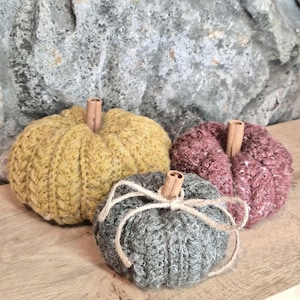 May include: Three handmade crocheted pumpkins in mustard yellow, dusty rose, and heather gray. Each pumpkin features a vertical ribbed texture and a natural cinnamon stick stem. The gray pumpkin is accented with a thin, light-colored twine bow.