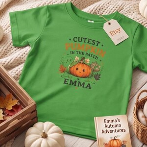 May include: A bright green short-sleeved t-shirt featuring a graphic of a smiling orange pumpkin surrounded by autumn leaves. The text reads "CUTEST PUMPKIN IN THE PATCH" and "EMMA" in dark green and orange lettering. An Etsy tag is attached to the collar.