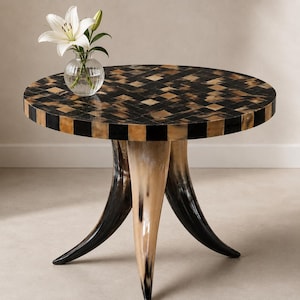 May include: A round table featuring a mosaic top with a black and tan checkered pattern. The table is supported by three curved legs made of horn-like material. A clear glass vase containing a white lily sits on the table surface.