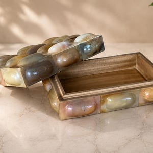 Puede incluir: Una caja rectangular de madera con una tapa que presenta una incrustación decorativa de piedras ovaladas lisas y pulidas en tonos bronceado, marrón y crema. La caja está abierta, revelando un interior de madera natural con acabado liso.