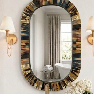Puede incluir: Un espejo de pared ovalado con un marco decorativo compuesto por segmentos de cuerno dispuestos verticalmente en tonos marrón, negro y crema. El espejo tiene una superficie reflectante lisa y un diseño de bordes redondeados.