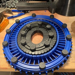 May include: A circular blue and black 3D printed knitting loom featuring a central ring with black pegs and an outer blue ring with numbered slots from 20 to 35. The device includes small black circular knobs positioned around the frame.