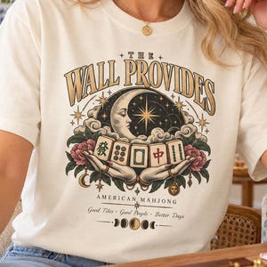 Può includere: Una t-shirt color crema con una stampa grafica che riporta il testo THE WALL PROVIDES e AMERICAN MAHJONG. Il design include una luna crescente, stelle, fiori e mani che tengono tessere del mahjong, con il testo Good Tiles, Good People, Better Days.