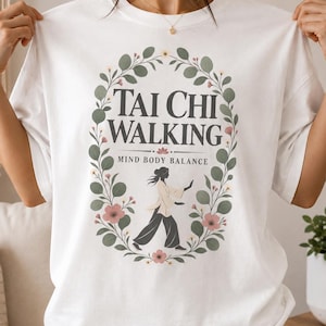 May include: A white t-shirt featuring a central graphic of a person practicing Tai Chi. The design includes the text "TAI CHI WALKING" and "MIND BODY BALANCE" surrounded by a decorative oval wreath of green leaves and small pink flowers.