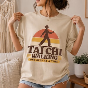 May include: A beige short-sleeve t-shirt featuring a graphic of a person practicing Tai Chi in front of a sunset. The text reads TAI CHI WALKING ONE STEP AT A TIME in dark brown letters. The design includes horizontal stripes in orange and yellow.