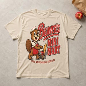May include: A beige t-shirt featuring a cartoon beaver wearing a red apron and a white hat, holding a shopping basket with a baguette and an apple. The text reads Beaver's Mini Mart and Your Neighborhood Favorite in red and white lettering.