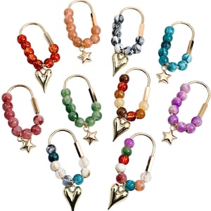 Puede incluir: Una colección de diez pendientes de aro dorados, cada uno con una hilera de cuentas de colores y un dije colgante. Los dijes consisten en un corazón dorado o una estrella dorada. Las cuentas varían en color, incluyendo rojo, naranja, verde, azul, morado y patrones jaspeados, creando un conjunto diverso de piezas de joyería.