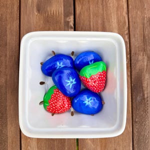 May include: A white square ceramic berry basket containing five hand-painted stones. Three stones are painted blue with white flower details, and two stones are painted red with green tops and white dots to resemble strawberries.