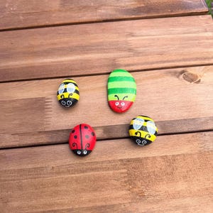May include: Four smooth, rounded stones painted as insects. Two stones are yellow and black striped bees with white wings, one is a red ladybird with black spots, and one is a green and yellow striped caterpillar with a red head and antennae.