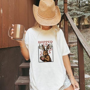 May include: A white t-shirt featuring a graphic of a jackalope holding a mug with steam rising, enclosed in a black square frame. Above the graphic, the text reads HOPPED UP in red letters. The shirt is worn with a tan straw cowboy hat.