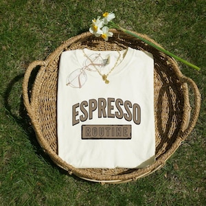 May include: A cream-colored t-shirt featuring the text ESPRESSO ROUTINE in brown block lettering. The shirt is displayed inside a round woven wicker basket alongside a pair of thin-framed glasses, a gold chain necklace, and a white flower.