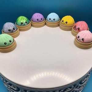 Può includere: Una collezione di nove piccole figure rotonde e colorate a forma di gnocco, ognuna con un volto sorridente con occhi neri e guance rosa. Ogni figura poggia su una base circolare color marrone chiaro, disposta a semicerchio.