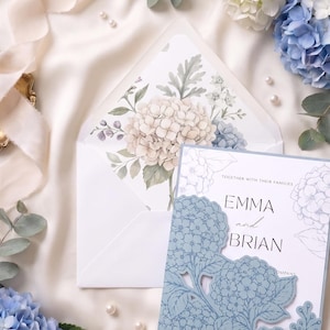 May include: A wedding invitation set featuring a white card with the text EMMA and BRIAN, enclosed in a dusty blue laser-cut cover with hydrangea motifs. The matching envelope has a floral hydrangea print on the inside flap.