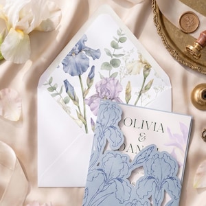 May include: A wedding invitation set featuring a light blue laser-cut card with an iris floral pattern, paired with an envelope lined with a watercolor iris and eucalyptus print. The invitation text reads Olivia & Daniel, with additional text below.