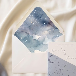 May include: A white envelope with a blue watercolor night sky liner featuring white stars, paired with a rectangular invitation card. The card has a light blue diagonal section with star constellations and a crescent moon, and the text "Bailey & Jason" and "Our love was written in the stars".