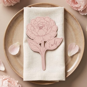 Könnte beinhalten: Ein hellrosa, gestanztes Pfingstrosen-Platzkärtchen mit dem Namen Emily in Schreibschrift auf einem der Blätter. Der florale Ausschnitt liegt auf einer gefalteten, cremefarbenen Leinen-Serviette, die auf einem runden Keramikteller platziert ist.