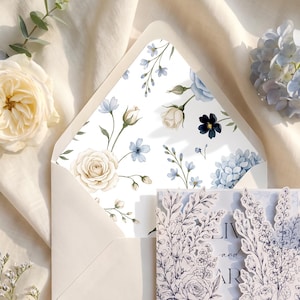 May include: A wedding invitation set featuring a cream envelope with a floral patterned liner in shades of blue and white. The invitation card has a laser-cut floral border in black ink on white paper, displaying text partially visible.