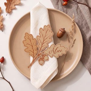 Peut inclure: Un marque-place en bois brun clair en forme de feuilles de chêne et de glands avec le prénom Alice gravé en écriture cursive. Le marque-place repose sur une serviette en tissu blanc pliée, posée dans une assiette en céramique beige lisse.