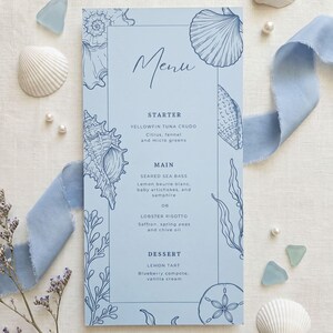 May include: A light blue rectangular menu card featuring dark blue line art illustrations of seashells, seaweed, and a sand dollar. The text lists a starter of yellowfin tuna crudo, main courses of seared sea bass or lobster risotto, and a lemon tart dessert.