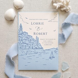 May include: A rectangular wedding invitation card featuring a light blue die-cut overlay of a coastal village and a sailboat. The cream-colored card displays the text: Together with their families, Lorrie & Robert request the pleasure of your company at their wedding celebration.