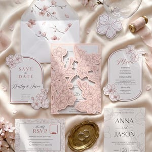 Peut inclure: Un ensemble de papeterie de mariage comprenant une carte Save the Date, une invitation découpée au laser avec des motifs de fleurs de cerisier, un menu et une carte RSVP. Les éléments sont imprimés en rose pâle et blanc avec des illustrations florales.