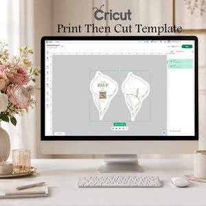 Puede incluir: Un monitor de computadora que muestra la interfaz del software Cricut Print Then Cut Template. La pantalla muestra dos dise&ntilde;os de tarjetas con forma de lirio de cala, uno con el texto RSVP y un c&oacute;digo QR. El monitor est&aacute; sobre un escritorio con teclado.