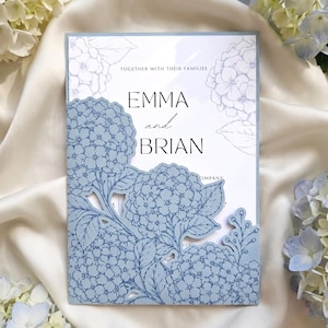 May include: A wedding invitation card featuring a dusty blue laser-cut cover with intricate hydrangea floral patterns. The white inner card displays the text TOGETHER WITH THEIR FAMILIES, EMMA and BRIAN, and partial text OMPANY.