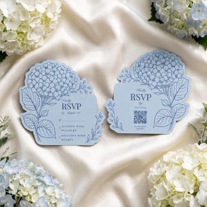 Puede incluir: Dos tarjetas de respuesta RSVP de color azul claro con formas troqueladas de hortensias. Las tarjetas presentan ilustraciones de líneas azul oscuro de flores y hojas. El texto incluye "Kindly RSVP by August 10", con un código QR en una tarjeta y una línea para nombres y casillas para "Accepts with pleasure" o "Declines with regret" en la otra.
