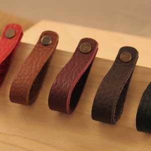 May include: Six leather drawer pulls in different colors, including red, brown, burgundy, dark brown, and black. Each pull has a small metal loop at the top.