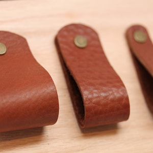 Op de afbeelding: Drie bruine lederen kastgrepen met messing accenten. De grepen zijn lusvormig en hebben een glad, getextureerd oppervlak.