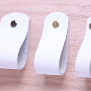 May include: Three white leather cabinet pulls with different coloured metal accents. The pulls are shaped like a rounded rectangle with a hole in the centre.