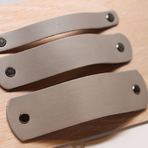 May include: Three gray leather cabinet pulls with silver metal hardware. The pulls are arranged in a stack, with the largest pull on the bottom and the smallest pull on the top.