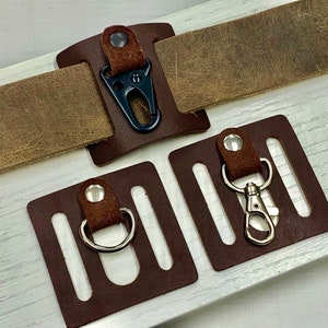 May include: Three brown leather keychains with metal hooks. The keychains have a rectangular shape with slots for holding keys. The keychains are attached to a brown leather strap.