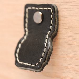 May include: Black leather handle with white stitching and a silver metal rivet. The handle is attached to a wooden surface.