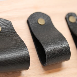 May include: Three black leather drawer pulls with brass hardware. The pulls are attached to a light wood surface.