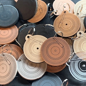 May include: A collection of leather and wood keychains shaped like vinyl records. The keychains feature a record player needle design and come in various colors, including black, brown, and beige.