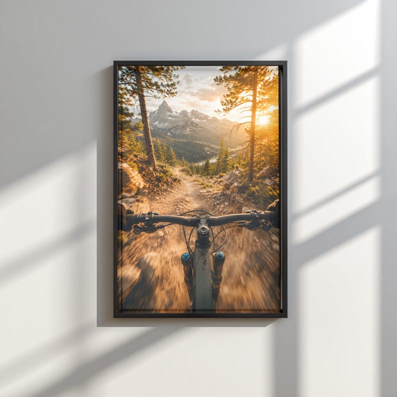 K&ouml;nnte beinhalten: Ein gerahmter Wandkunstdruck mit einer Ego-Perspektive beim Fahren eines Mountainbikes auf einem Feldweg. Die Szene zeigt hohe Kiefern, einen steinigen Pfad und schneebedeckte Berge in der Ferne unter einem hellen, goldenen Sonnenuntergangshimmel.