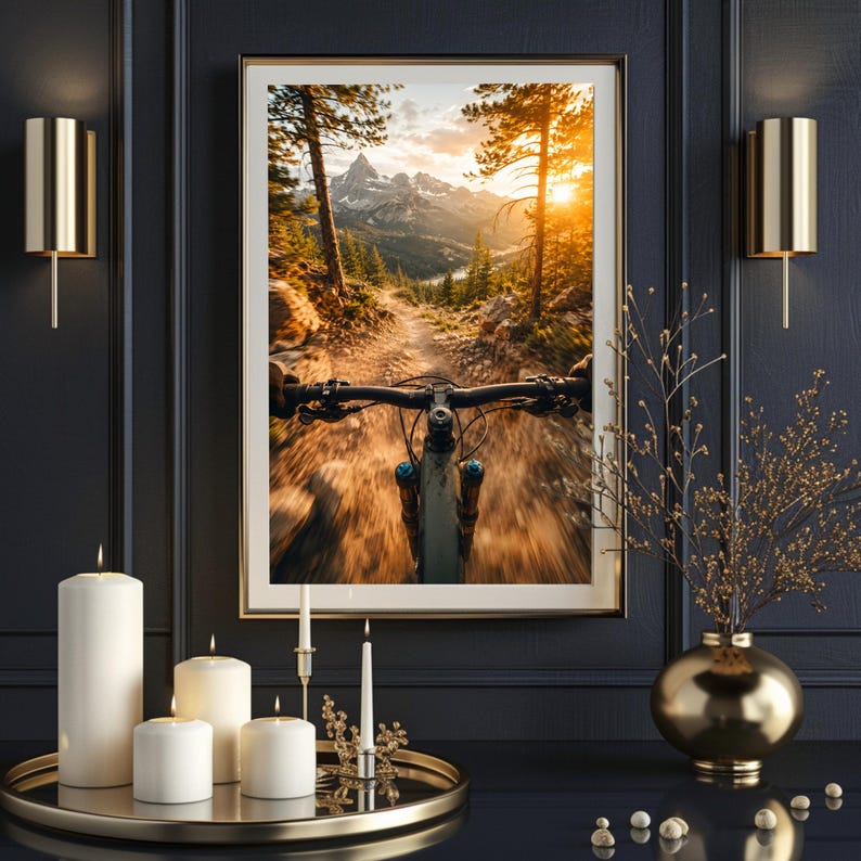 K&ouml;nnte beinhalten: Ein gerahmter Wandkunstdruck mit einer Ich-Perspektive eines Mountainbikes auf einem Feldweg, umgeben von Kiefern und Bergen bei Sonnenuntergang. Das Bild h&auml;ngt an einer dunklen Wand zwischen zwei goldenen Wandleuchten, mit Kerzen auf einem Tablett.