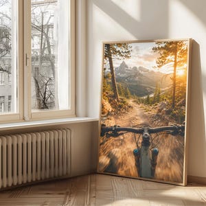 K&ouml;nnte beinhalten: Ein gerahmter Wandkunstdruck, der die Ich-Perspektive beim Mountainbiken auf einem Waldweg zeigt. Die Szene umfasst hohe Kiefern, einen steinigen Pfad und ferne schneebedeckte Berge unter einem hellen, goldenen Sonnenuntergang.
