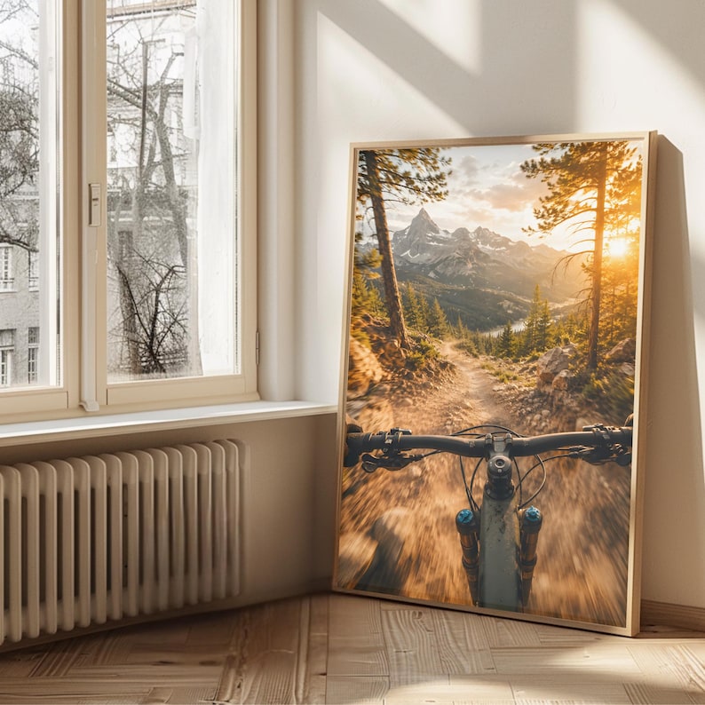 K&ouml;nnte beinhalten: Ein gerahmter Wandkunstdruck, der die Ich-Perspektive beim Mountainbiken auf einem Waldweg zeigt. Die Szene umfasst hohe Kiefern, einen steinigen Pfad und ferne schneebedeckte Berge unter einem hellen, goldenen Sonnenuntergang.