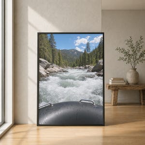 Può includere: Una stampa artistica incorniciata che mostra una vista in prima persona da un gommone nero che naviga su un fiume impetuoso. La scena presenta rapide di acqua bianca, grandi massi grigi, fitti pini verdi e un cielo azzurro con nuvole.
