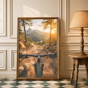 K&ouml;nnte beinhalten: Ein gerahmter Kunstdruck mit einer Ich-Perspektive auf einen Mountainbike-Lenker und eine Federgabel auf einem Feldweg. Die Szene zeigt einen sonnendurchfluteten Waldweg, der w&auml;hrend eines goldenen Sonnenuntergangs zu Berggipfeln f&uuml;hrt.