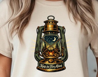 May include: A cream-colored short-sleeve t-shirt featuring a graphic of a vintage-style lantern. Inside the lantern glass is a night scene with a campfire, pine trees, and a crescent moon. The text "Keep the Fire Alive" is printed at the base.