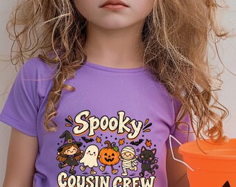 May include: A light purple t-shirt featuring a graphic of a witch, ghost, pumpkin, mummy, and black cat with the text Spooky Cousin Crew. The shirt is paired with a bright orange plastic jack-o'-lantern bucket and purple sunglasses.
