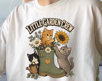 May include: A cream-colored t-shirt featuring a graphic print of three kittens and a green rain boot. The text "LITTLE GARDEN CREW" is arched above the illustration, which includes sunflowers, daisies, and a small watering can in a garden setting.